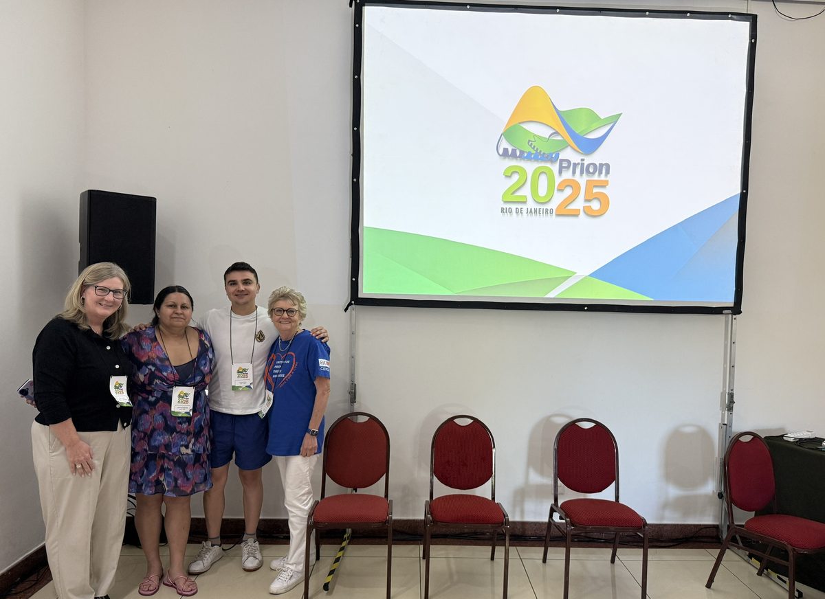 FEN en la conferencia Prion 2025: cuatro asistentes con credencial ante la pantalla con el logotipo del congreso.